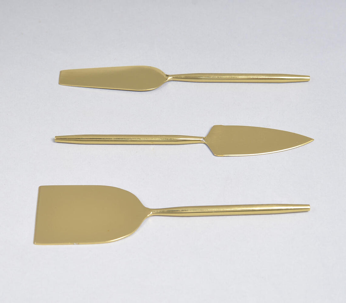 A set of three gold-colored stainless steel cheese serving utensils, including a cheese knife, a cheese spreader, and a cheese wedge server, displayed against a white background.