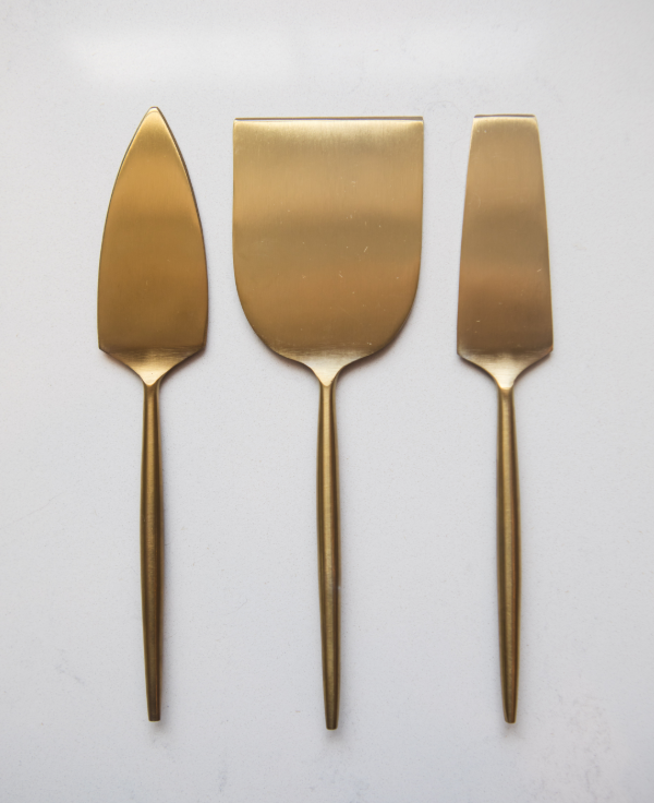 A set of three gold-colored stainless steel cheese serving utensils, including a cheese knife, a cheese spreader, and a cheese wedge server, displayed against a white background.