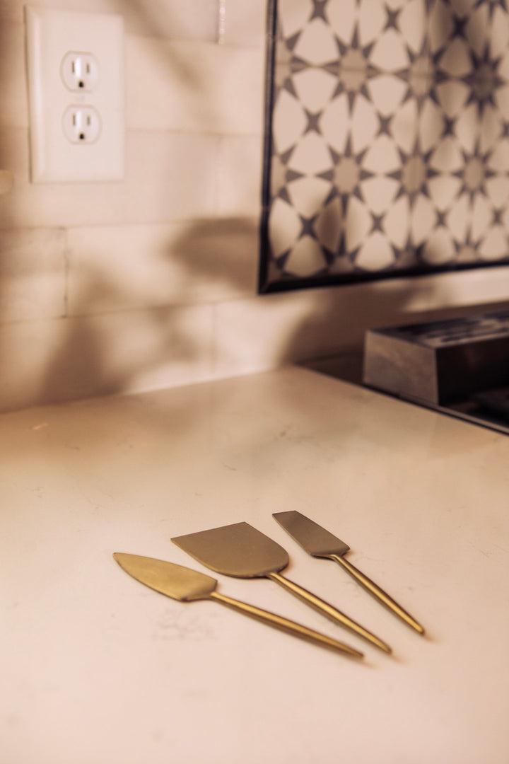 Three gold spatulas on a light-colored countertop with a decorative wall tile in the background.